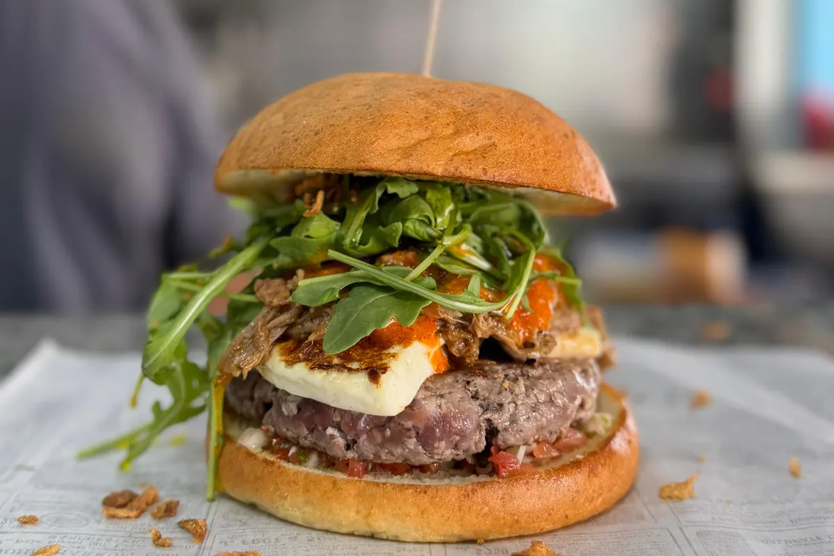 Una hamburguesa reci&eacute;n montada sobre papel: pan brioche, r&uacute;cula, queso a la plancha y cebolla crujiente, con la cocina desenfocada detr&aacute;s