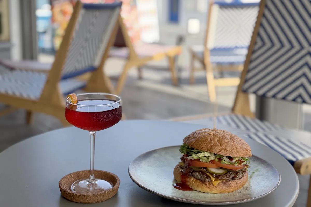 Una hamburguesa y un c&oacute;ctel rojo en una mesa de la terraza, con tumbonas y el mar al fondo