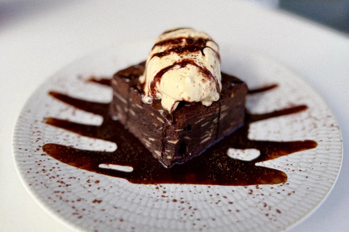 Un brownie de chocolate con una bola de helado y sirope, servido en un plato blanco espolvoreado con cacao