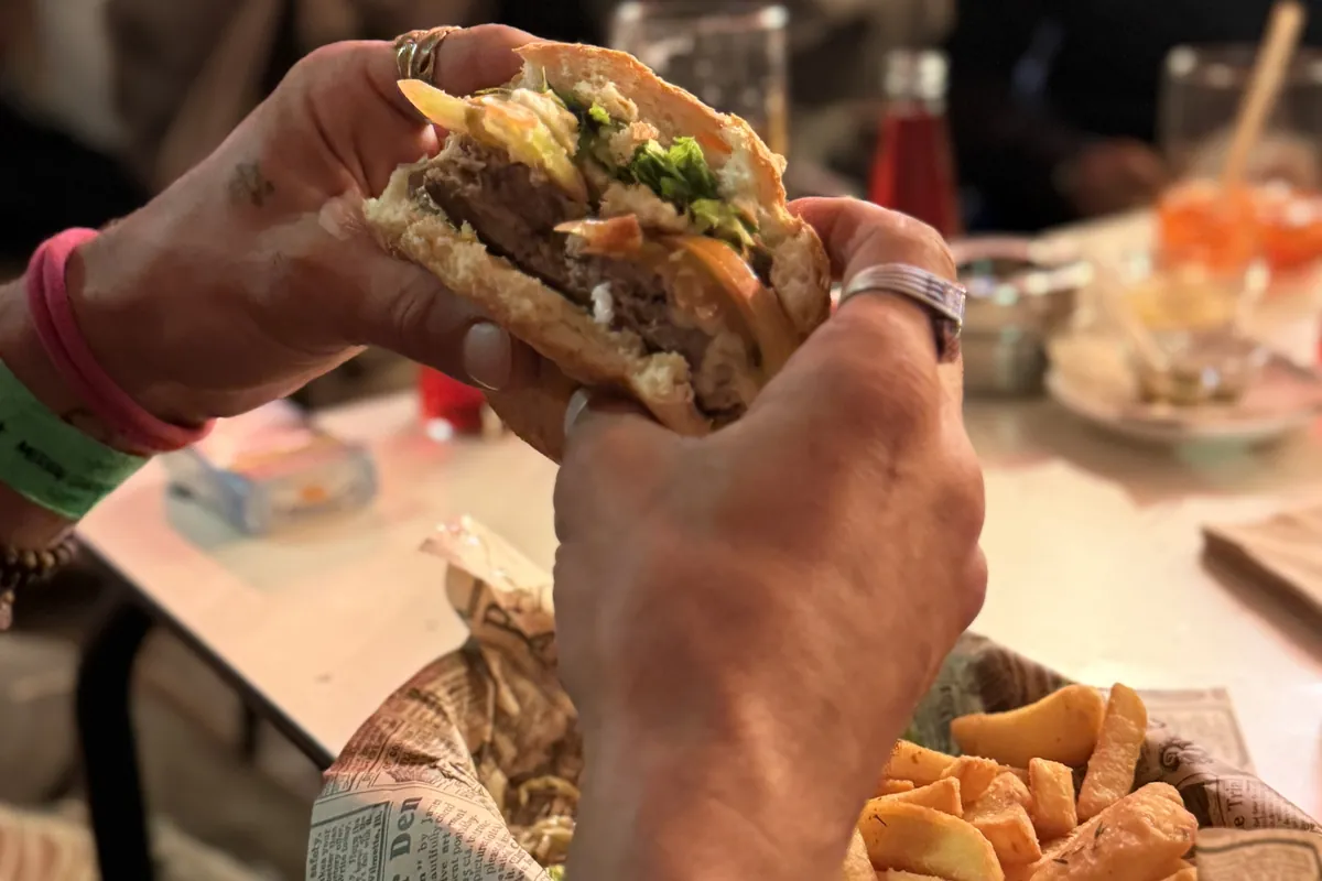 Un cliente sujeta una hamburguesa a medio comer sobre una cesta de patatas, en una mesa llena de gente