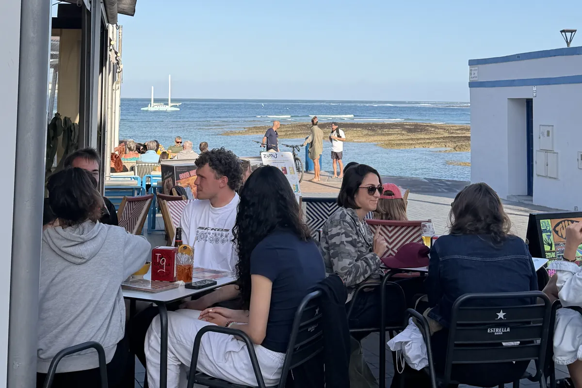 Mesas de la terraza con clientes y, justo enfrente, el mar con un velero y la orilla