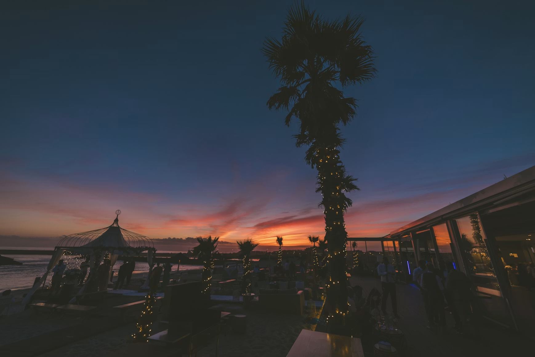 Un bar de playa a la hora dorada: una palmera con luces, mesas en la terraza y el mar bajo un cielo rosa y naranja de puesta de sol