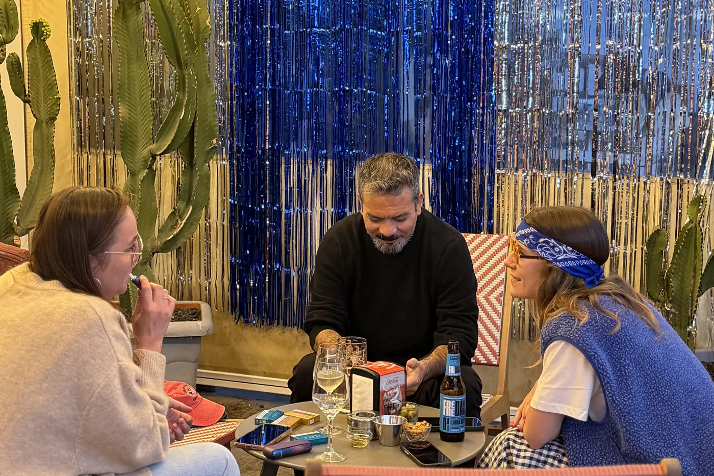 Tres personas charlando con sus copas en una mesa de Twinny, con la cortina de tiras azul y los cactus de fondo